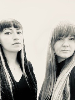 Boyd & Grogan - Artist Duo. Portrait photograph. Black and white portrait of the artists. The artists are stood together, both looking towards the camera. They are both dressed in black and have long straight hair with fringes.
