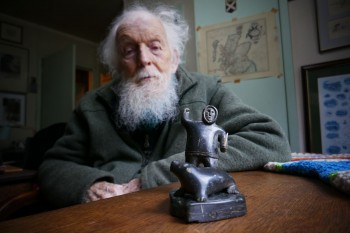 Photograph of Adam Watson, with a stone and ivory carving of a hunter and seal Photograph of Adam Watson later in life. On a table in front of him is a stone and ivory carving of a hunter and seal