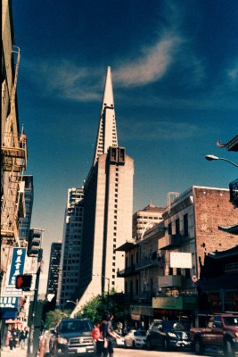 Transamerica Pyramid, San Francisco photograph of a street, the sky is blue and there is a pyramid shaped bulding in the centre of the image, surrounded by other building. People and cars can be seen in the foreground on the street.