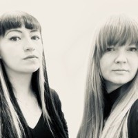 Black and white portrait of the artists.  The artists are stood together, both looking towards the camera.  They are both dressed in black and have long straight hair with fringes. 